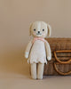 A Cuddle + Kind Dog Stuffed Animal with a smiling face, wearing a pastel pink scarf, stands upright next to a woven basket on a light brown background.