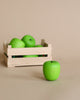 A wooden crate filled with Erzi Apple Green Pretend Food miniature green apples on a beige background, with one apple placed in front of the crate.