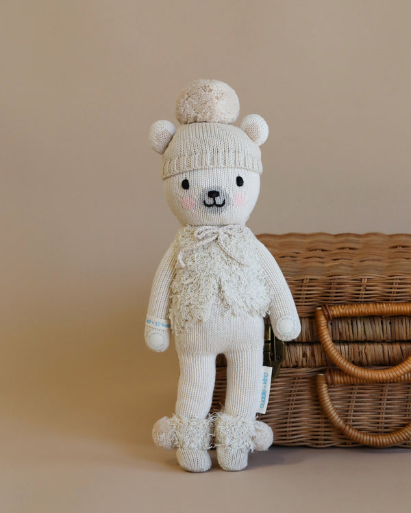 The Cuddle + Kind Stella The Polar Bear, a hand-knit plush bear, stands upright beside a wicker basket. It is wearing a beanie adorned with a pom-pom and matching textured scarf and booties. Made with hypoallergenic materials, the bear has a smiling face with rosy cheeks and small round ears, all in a beige and white color palette.