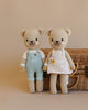 Two Cuddle + Kind the Honey Bears, one in blue overalls and the other in a white dress with flowers, standing against a wicker basket on a beige background.