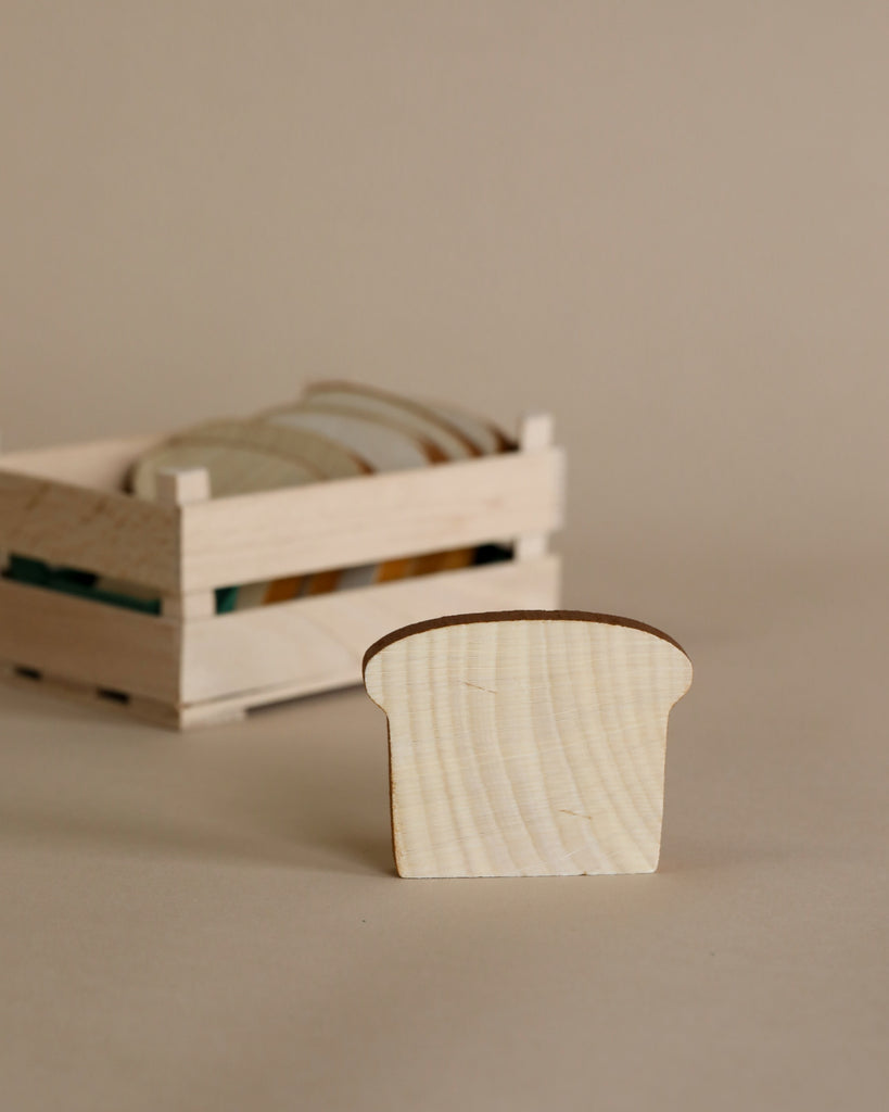 A small, pale wooden Erzi Slice of Toast Pretend Food handcrafted toy in the foreground with a crate of similar objects blurred in the background, all against a plain beige backdrop.