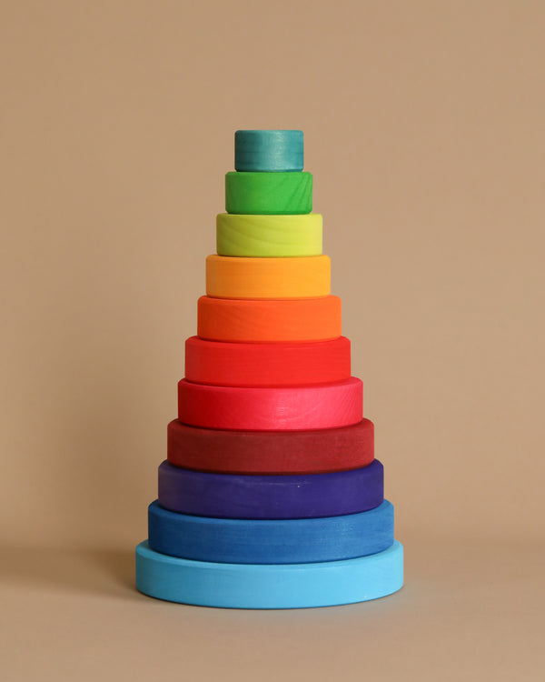 The Grimm's Stacking Tower is a captivating wooden toy featuring rainbow-colored circular rings arranged in descending size order from blue at the bottom to green on top, resembling a conical tower. It not only delights with its vibrant colors but also promotes fine motor skills and eye-hand coordination. The toy is set against a plain beige background.