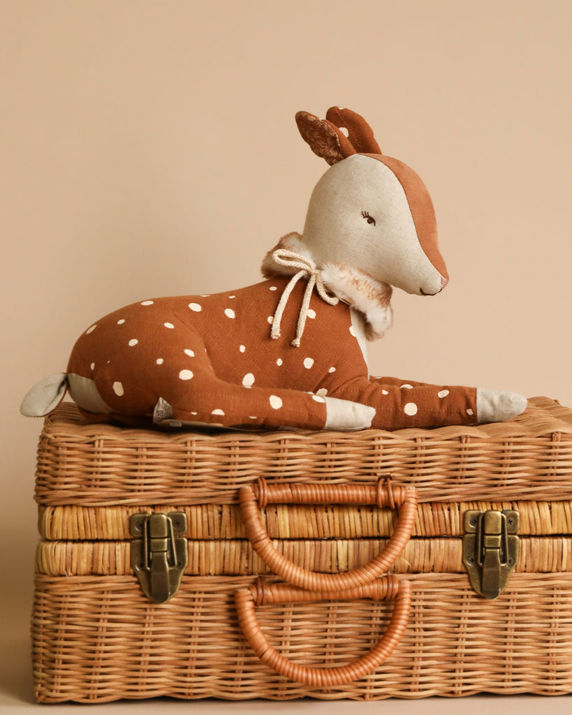 A plush Maileg Cosy Bambi Deer - Off white, resembling a brown fawn with white spots and a white face, is lying on top of a closed, woven wicker suitcase. The classic item features two leather straps securing the front and two handles. The background is a plain beige color, complementing the cotton linen aesthetic.