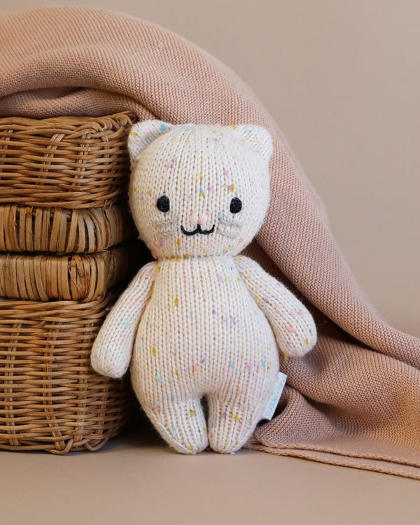 A hand-knit Cuddle + Kind Baby Kitten made from Peruvian cotton yarn, with multicolored specks, sitting beside a wicker basket draped with a soft pink cloth, against a beige background.