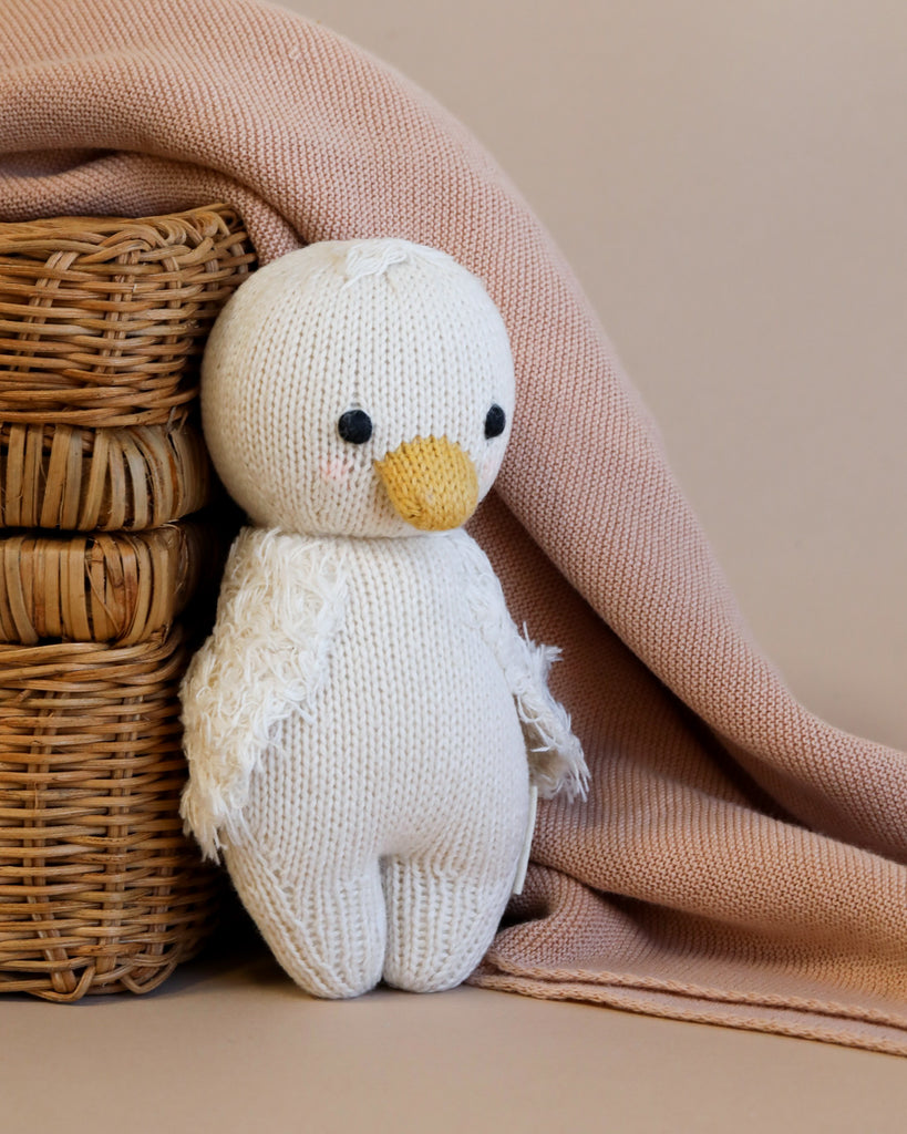 A hand-knit Cuddle + Kind Baby Gosling plush toy with a yellow beak and black eyes, nestled against a soft pink blanket next to a small wicker basket.