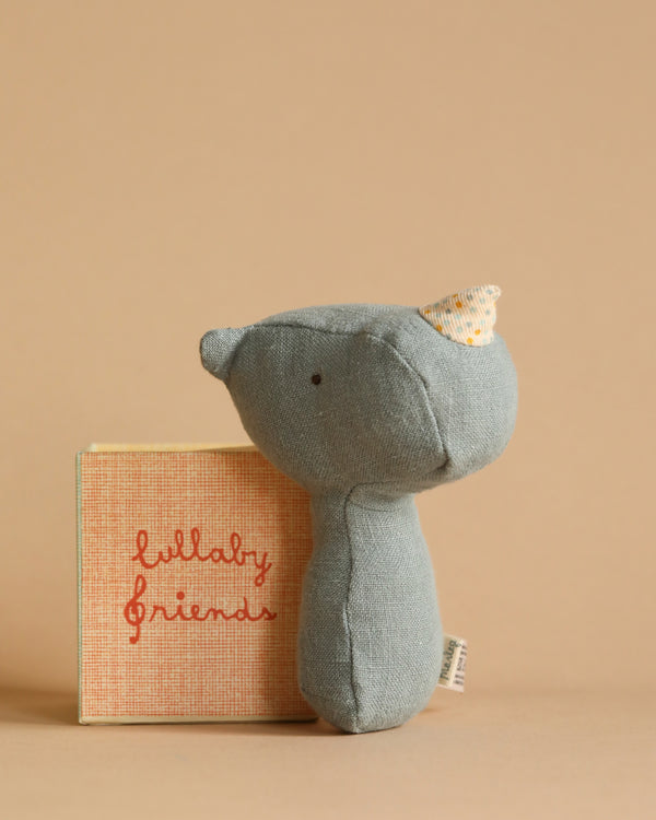 A soft, dusty blue stuffed animal toy resembling a rhino from the Maileg Lullaby Friend Rattles collection stands next to a small orange and beige book with "lullaby friends" written on the cover. The background is a solid light brown.