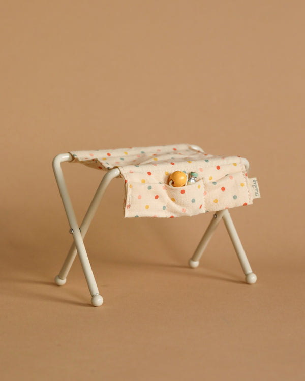 The Maileg Nursery Table, Baby Mouse - Off White is a compact, foldable stool featuring a metal frame and covered with beige fabric adorned with colorful dots. The fabric includes two front pockets, housing a tiny doll and a small toy bottle. It has the appearance of a nursery table, seamlessly blending into its smooth tan backdrop.