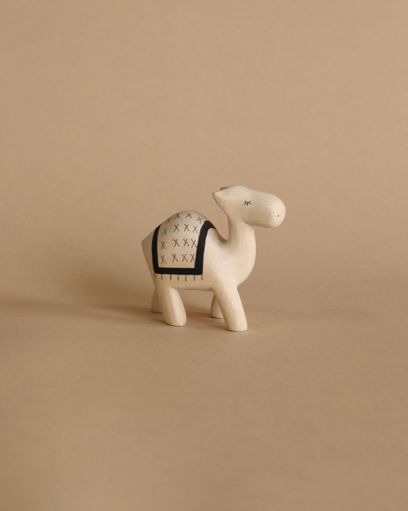 A small, minimalist Handmade Wooden Camel with abstract black patterns stands on a beige background. The figurine, reminiscent of handcrafted wooden animals, has a rectangular patterned cloth depicted on its back and is designed in a simplified, stylized form.