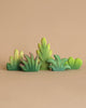 The Bumbu Wooden Bushes Set features wooden toy plants in green shades arranged against a plain beige background. The intricate carvings depict various leaf shapes and sizes, ranging from tall and thin to wide and bushy, beautifully illustrating the diversity of plant life through simple, stylized designs. This handmade set is perfect for eco-friendly play.