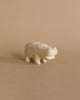 The Handmade Tiny Wooden Safari Animals - Hippo is a small, light-colored figurine crafted from Albizia wood, standing on a plain, beige background. This minimalist handcrafted piece features a rounded body, short legs, and a wide, smiling mouth.