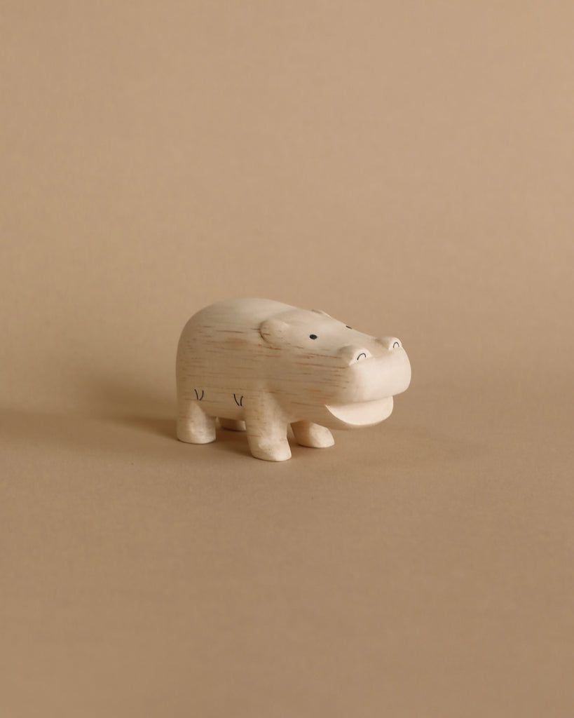 The Handmade Tiny Wooden Safari Animals - Hippo is a small, light-colored figurine crafted from Albizia wood, standing on a plain, beige background. This minimalist handcrafted piece features a rounded body, short legs, and a wide, smiling mouth.