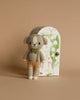 A small, hand-knit cotton doll of a stuffed dog named "Cuddle + Kind Tiny Noah The Dog," dressed in beige overalls and a green bow tie, stands in front of a whimsical, semi-circular box adorned with floral illustrations and the text "Fleur the dog." This fair trade handmade treasure is set against a solid, warm beige background.