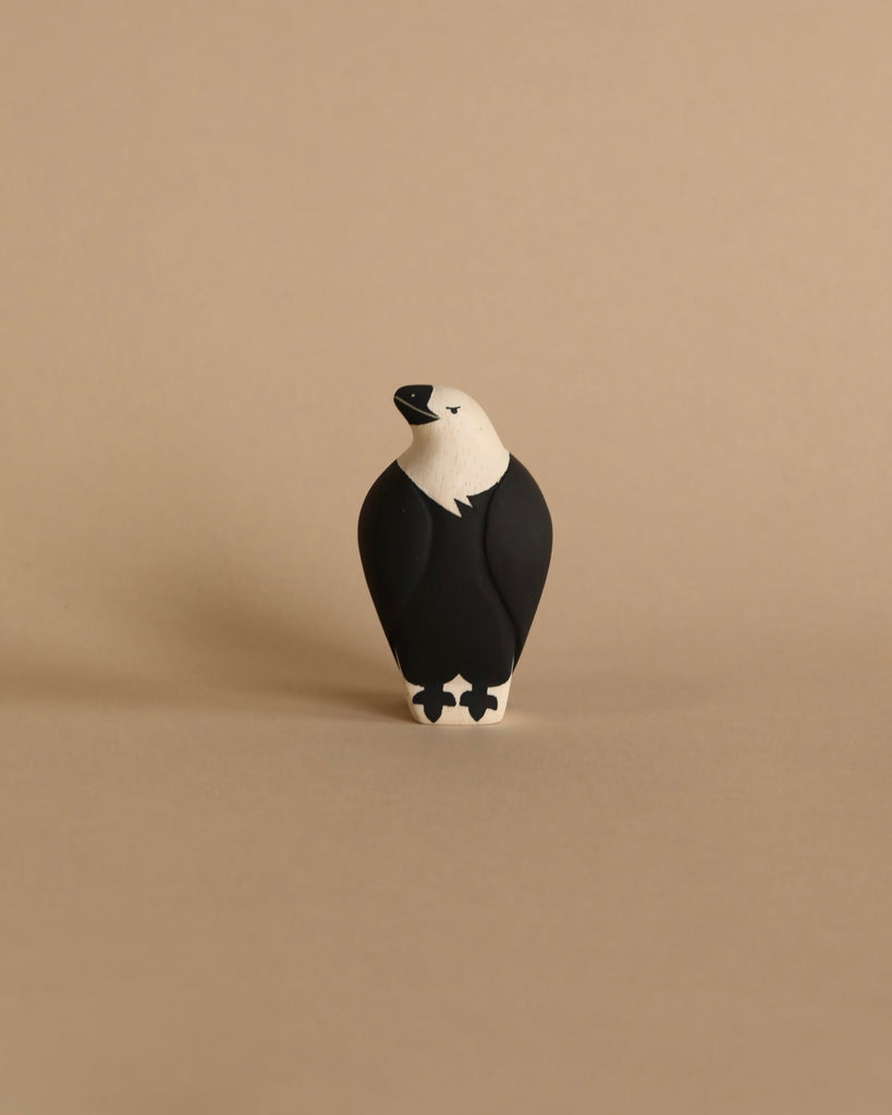 A small, minimalist figurine of a black and white bird, part of the "Handmade Tiny Wooden Forest Animals - Eagle" collection, stands against a plain beige background. Handcrafted by skilled artisans from Albizia wood, the eagle has a white head and beak with a black body and legs. The artistic style is simple and clean.