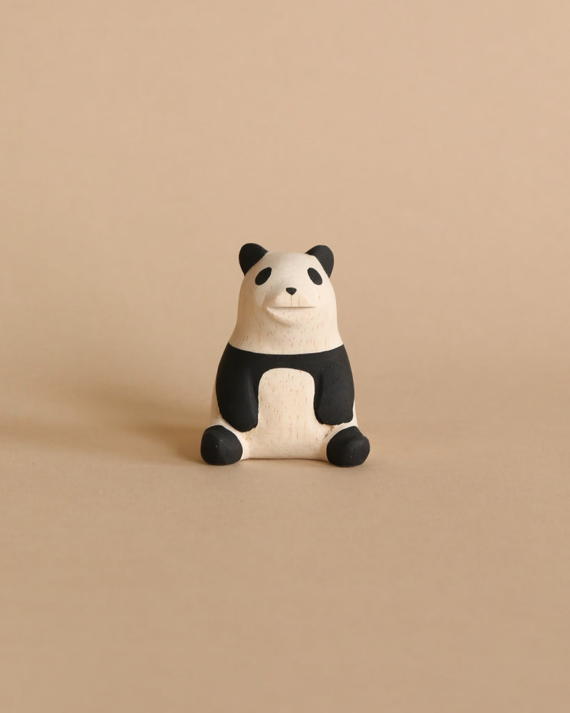 The Handmade Tiny Wooden Panda, meticulously crafted from Albizia wood, is set against a plain beige background. This charming figurine features a smiling expression, black ears, black patches around its eyes, and black limbs. The rest of its body is painted white.