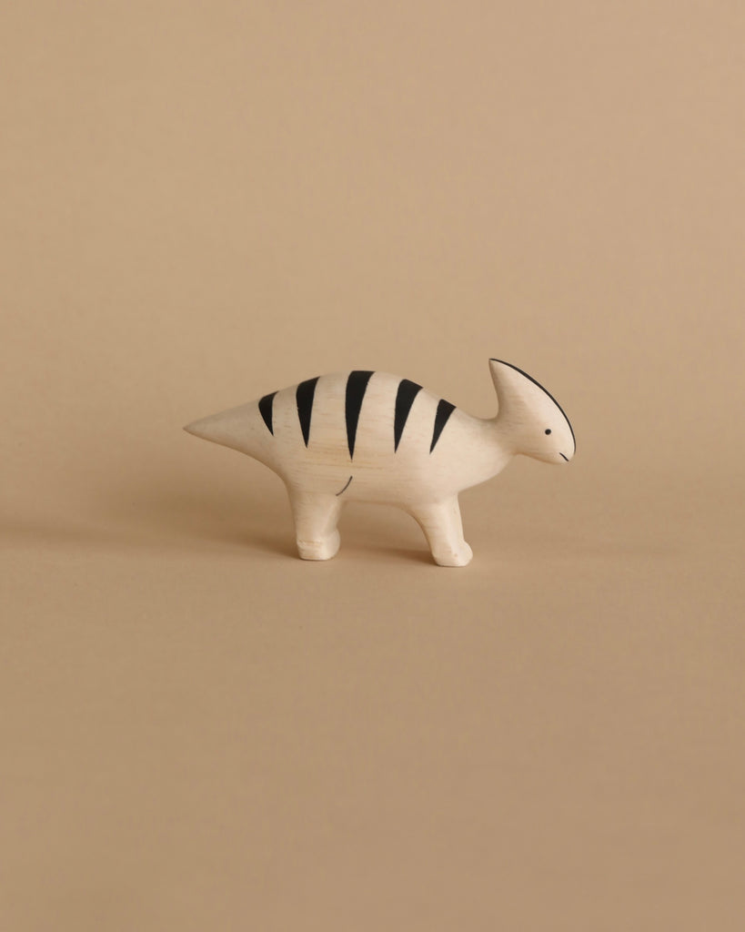 The Handmade Wooden Parasaurolophus, a minimalist and handmade toy, is crafted in the shape of a small dinosaur with a cream-colored body and black stripes. It stands against a beige background, featuring simple, stylized details that give it a modern yet playful look. Made from Albizia wood, this handcrafted wooden animal embodies both charm and elegance.