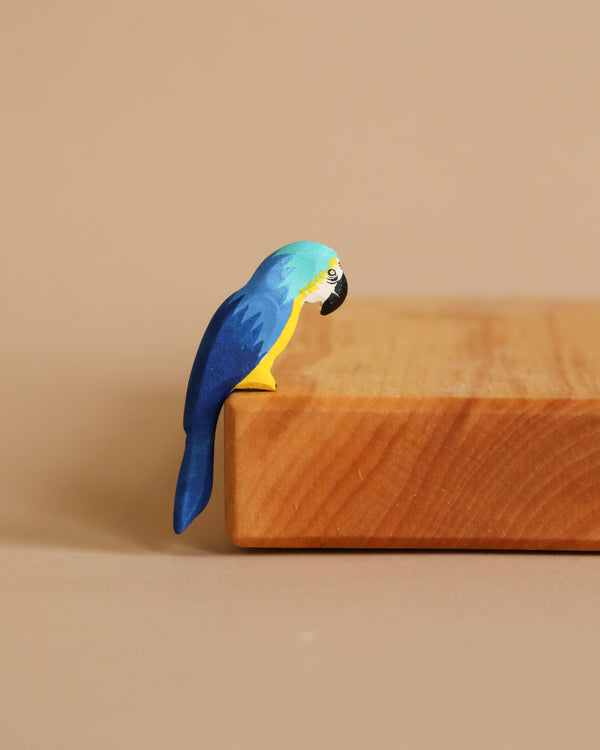 The Bumbu Wooden Blue Macaw Parrot perches on the edge of a wooden surface against a beige background. Made from eco-friendly materials, this small, colorful figurine features detailed craftsmanship that lends it a lifelike appearance.