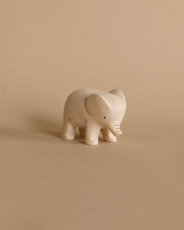 A small **Handmade Tiny Wooden Safari Animals - Elephant** with a smooth, light-colored finish stands on a beige background. Made from Albizia wood, the elephant features simple, carved details for its eyes and trunk.