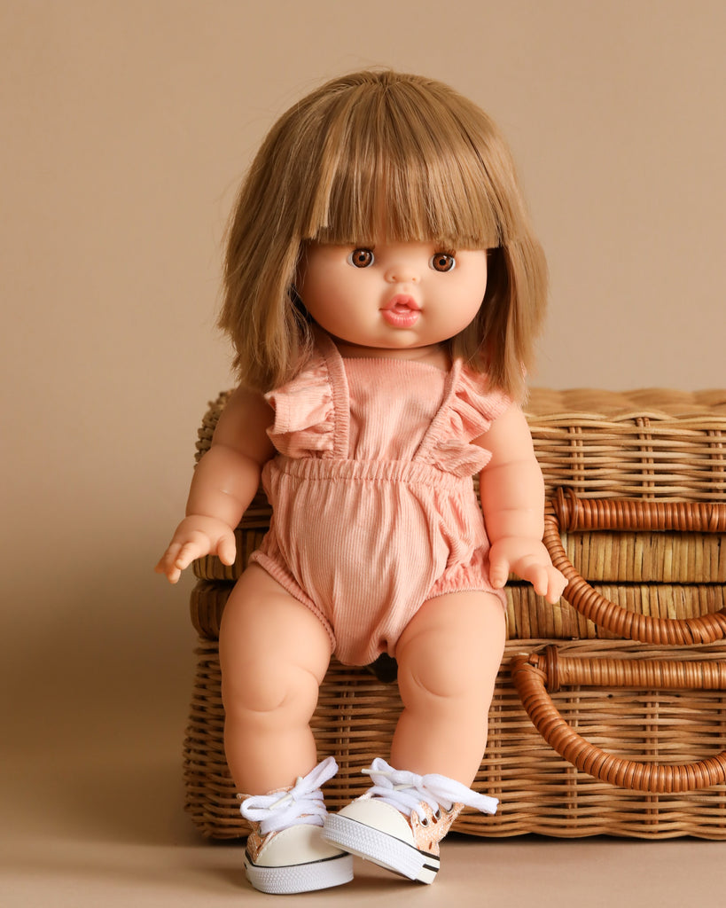 A Minikane Baby Doll (13") - Zoe, characterized by her light brown hair with straight bangs and brown eyes, stands next to a wicker basket. Zoe is dressed in a light pink sleeveless romper with ruffled straps and white sneakers featuring pink accents and white shoelaces. She exudes a natural vanilla scent.