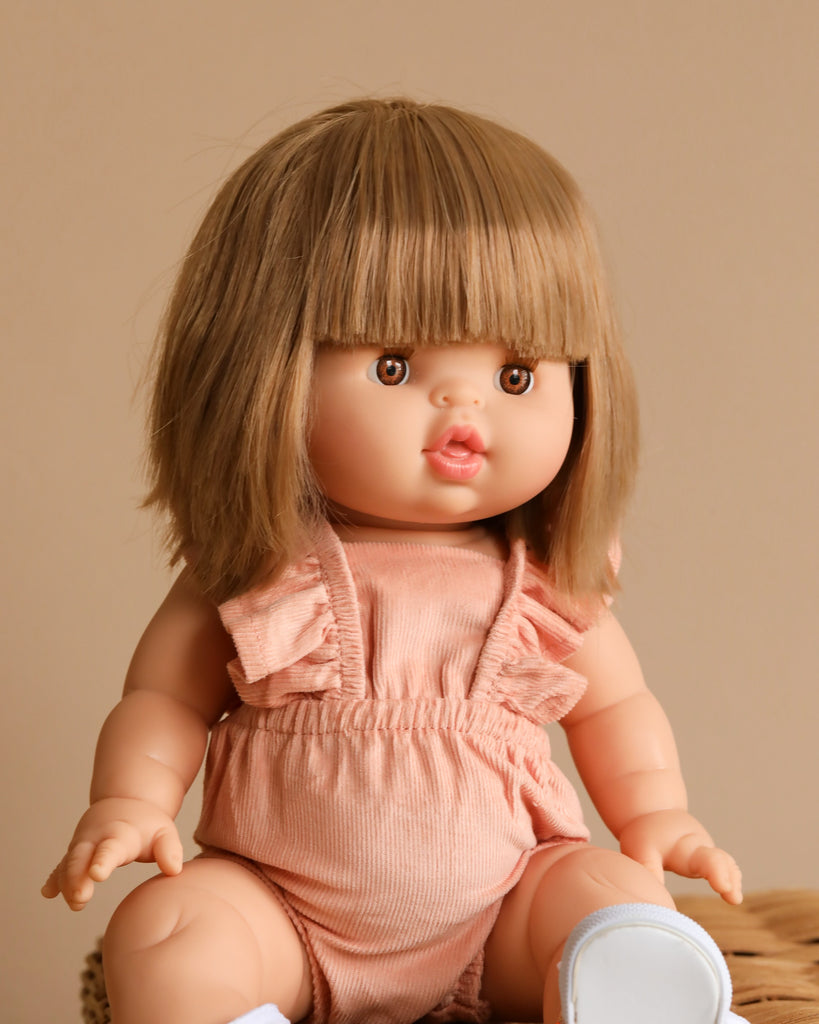 A close-up of the Minikane Baby Doll (13") - Zoe with short, straight, light brown hair and bangs. The doll is dressed in a pink romper with ruffled sleeves and white shoes. Emitting a natural vanilla scent, the background is a neutral beige color.