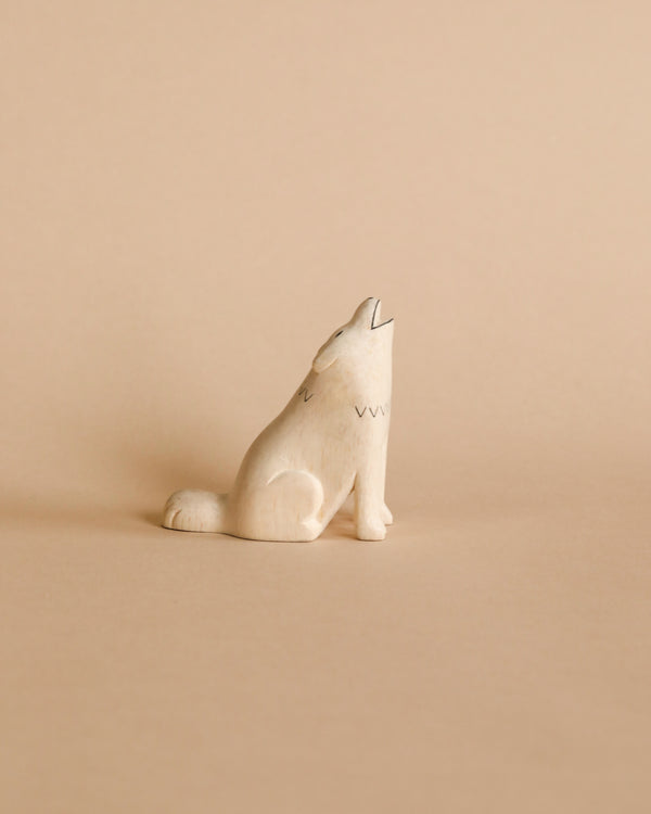 The Handmade Wooden Wolf, a small white wooden figurine of a howling wolf, rests on a beige surface set against a light brown background. Expertly handcrafted by Bali artisans from Albizia wood, this minimalist sculpture features simple etched details for the eyes, nose, and fur, lending it a clean and modern appearance.