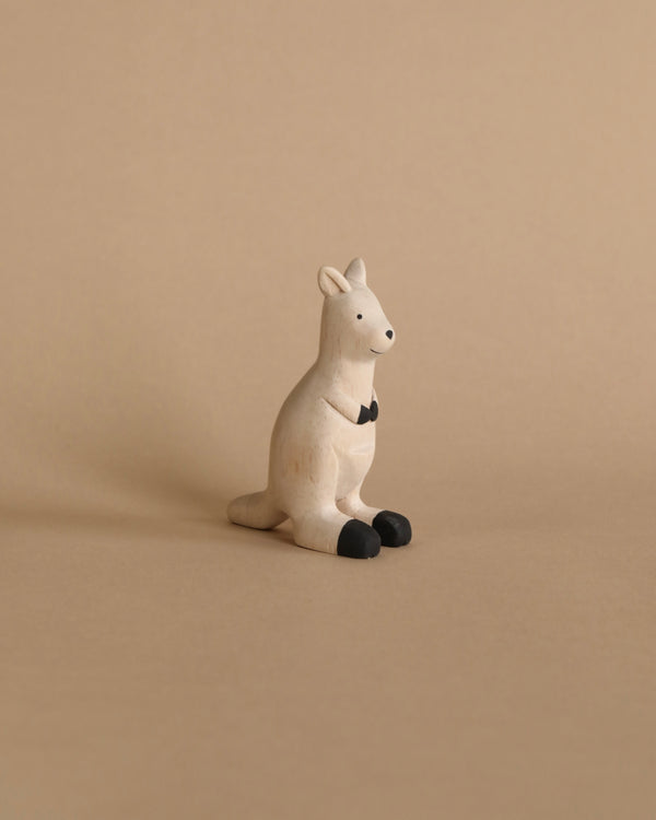 A small Handmade Wooden Kangaroo figurine, painted white with a black bowtie and black feet, is seated on a light brown surface. Handcrafted wooden animals like this are often created by Bali artisan products. The background is the same light brown color, creating a neutral and cohesive backdrop.