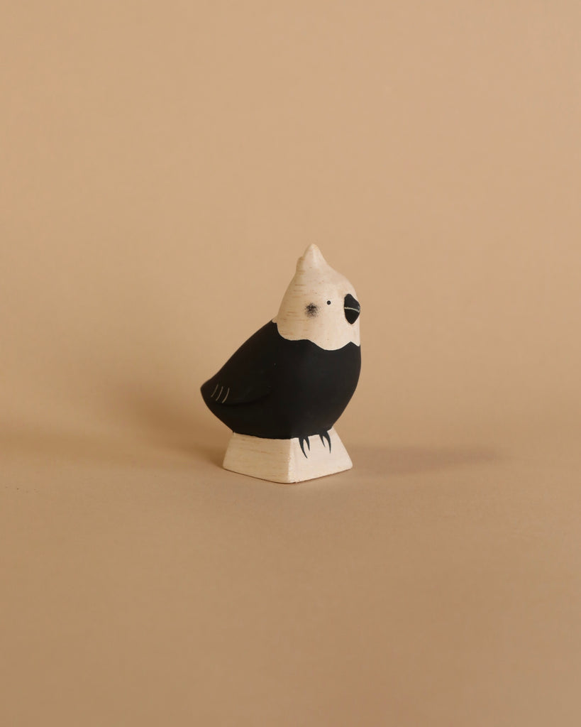 The Handmade Wooden Parrot stands against a plain beige background, showcasing its small, decorative charm. Handcrafted from Albizia wood, the parrot features a black body contrasted by a white head, beak, and base for a striking two-toned appearance. Its minimal detailing adds to its simple and elegant look.