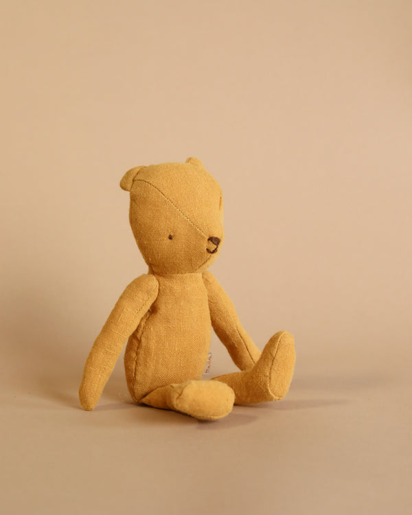 A Maileg Teddy Junior made of soft linen is sitting upright against a plain beige background. The bear, with its vintage look, has simple stitching for its eyes and nose, and its limbs are slightly spread out. The overall appearance is minimalist and handcrafted.