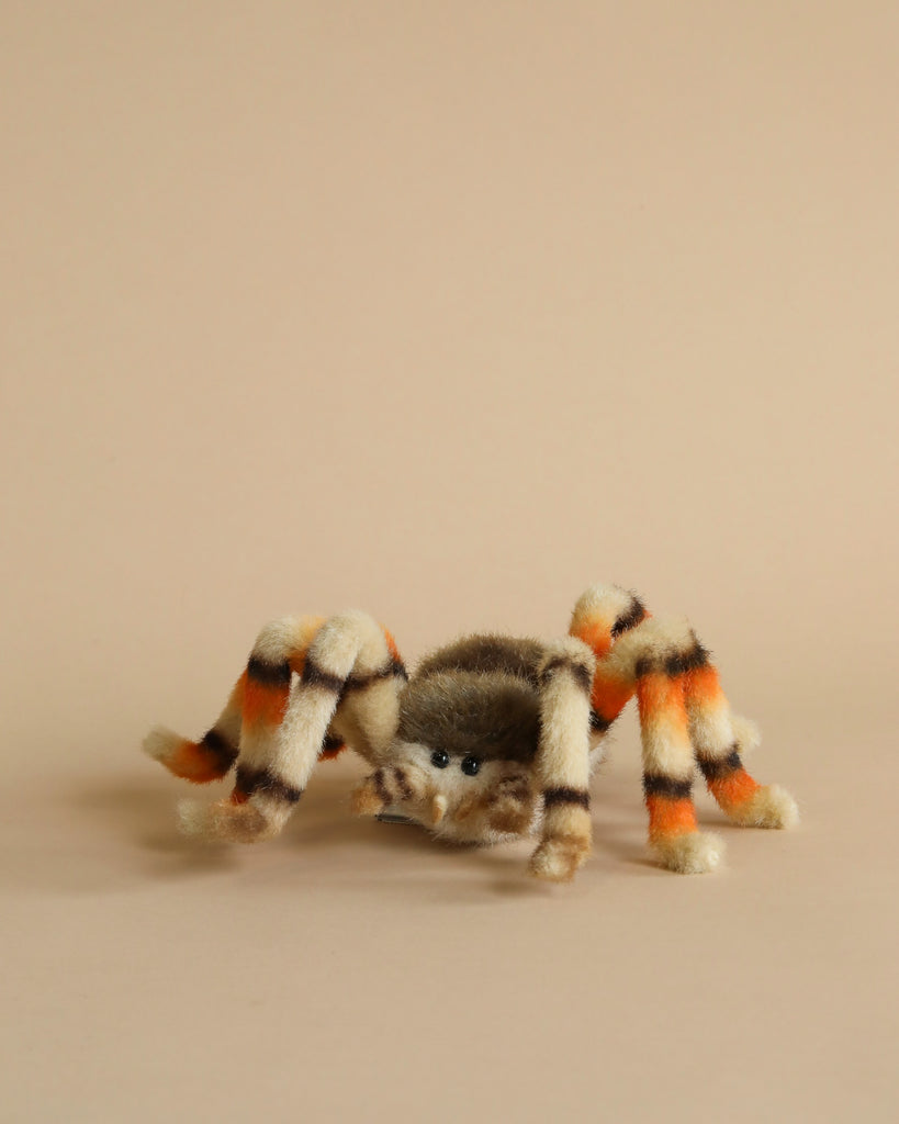 A Jumping Spider Stuffed Animal with a fuzzy texture, featuring an orange, black, and cream striped pattern. This unique handcrafted toy has large black eyes and is set against a plain beige background.