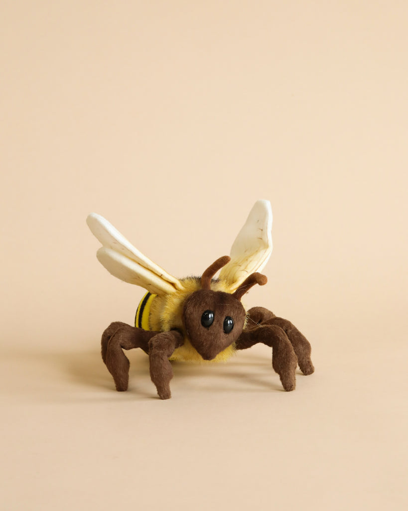 A Honey Bee Stuffed Animal, featuring a yellow and black striped body, brown legs, white wings, and large black eyes, is set against a plain beige background. This hand-sewn animal is detailed and lifelike, with visible textures on its body and wings.