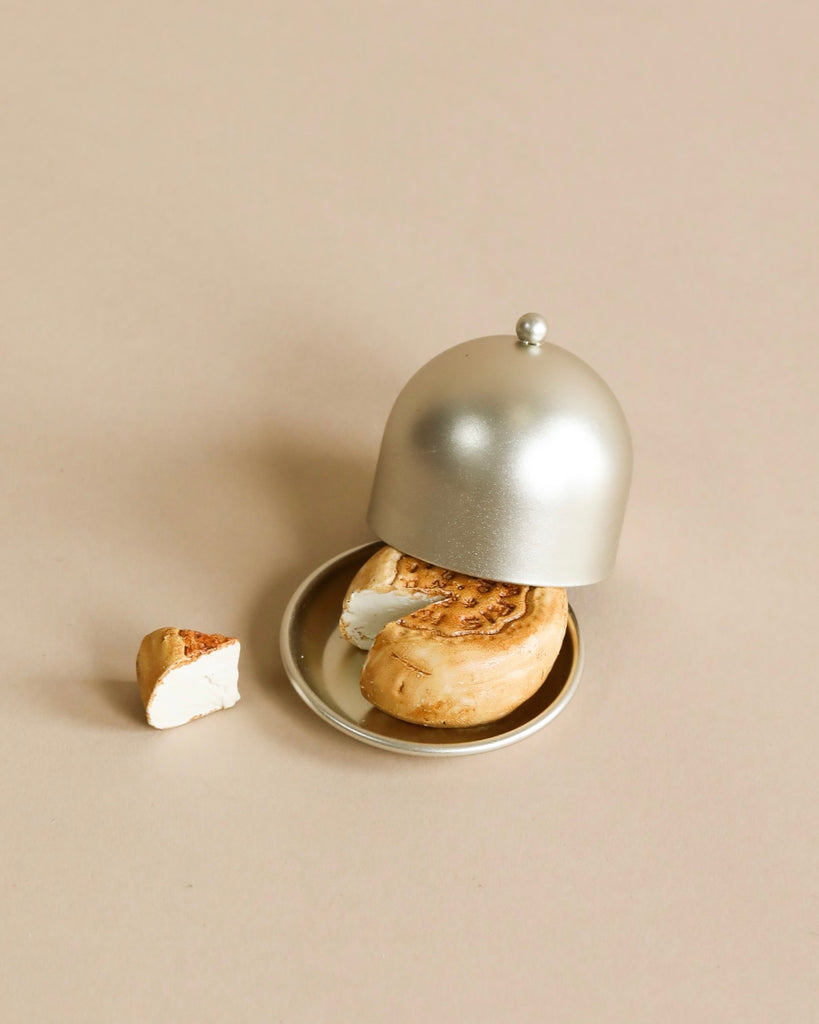 A partially eaten Maileg Cheese Bell, a delicious surprise, rests on a small plate with a silver cloche cover. A piece of the sweet treat lies beside the plate on a light beige surface, almost as if tempting someone—perhaps even the household mice—to indulge.