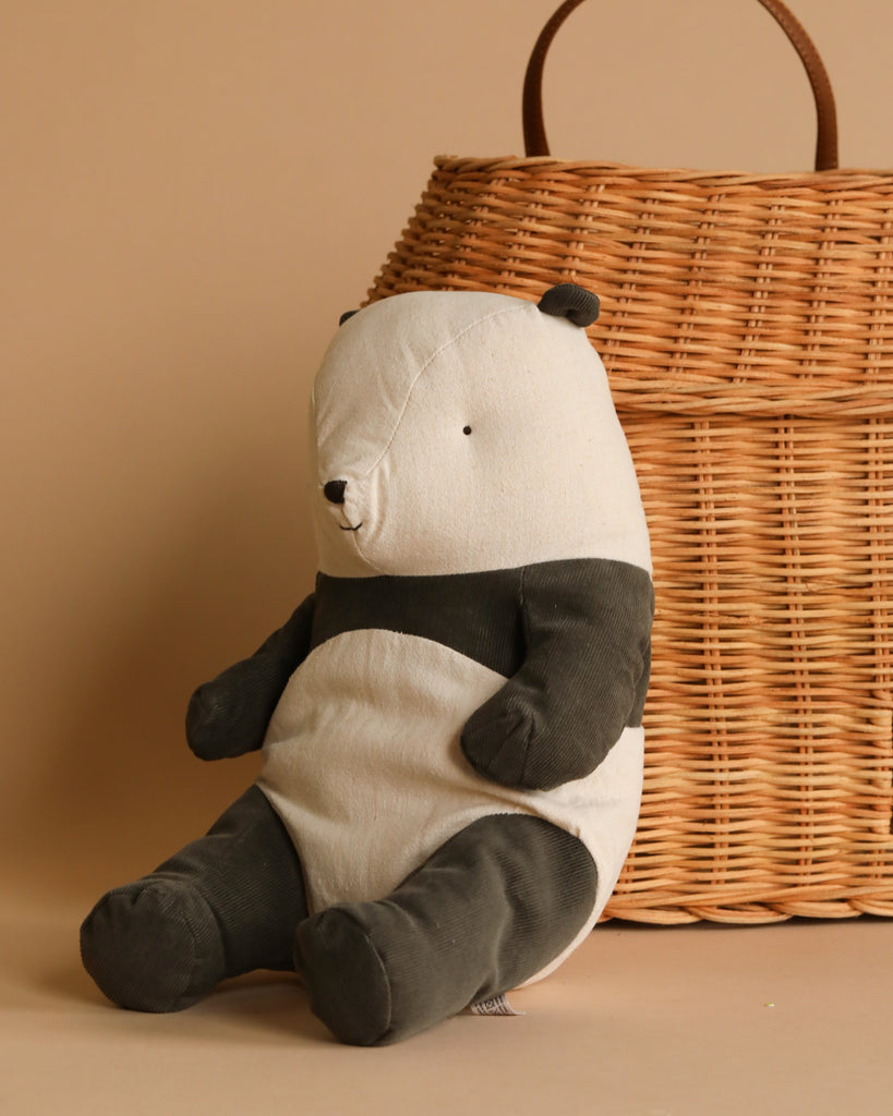 A Maileg Panda Stuffed Animal, Medium, with black and white features sits leaning against a large wicker basket. The background is a muted beige, creating a warm and cozy ambiance. Part of the Maileg Safari friends collection, this plush toy is crafted from recycled polyester for an eco-friendly touch.