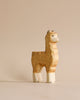 A Handmade Holzwald Alpaca, crafted with natural dyes, exhibits a cheerful expression while standing upright against a plain, light beige background.