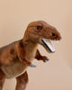 A realistic T-Rex Stuffed Animal stands against a plain beige background. This hand-sewn animal features brown fur, an open mouth with visible white teeth, and small, gray forelimbs. The dinosaur is depicted in a dynamic, roaring pose, showcasing its unique features.