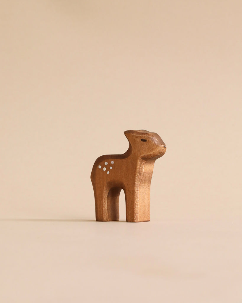 A Handmade Holzwald Standing Fawn figurine with dotted spots and pricked ears stands against a plain, light beige background, representing a delightful example of sustainable toys.