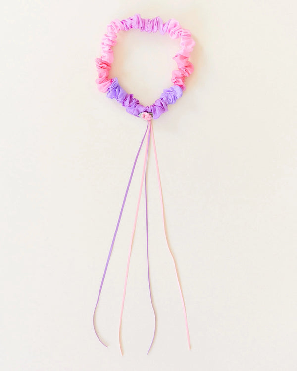 A Sarah's Silk Blossom Garland Headband is pink and purple with long dangling ribbons, reminiscent of festive garlands. The headband is ruched and the ribbons extend from the bottom, adding a decorative touch. Perfect for dress-up play, it is set against a plain, light background.