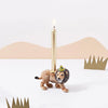 A unique Lion Cake Topper shaped like a lion, crafted from fine porcelain, featuring a lit slender taper candle on its back, positioned against a minimalist background with abstract brown mountains and green cutouts.