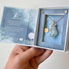 A hand holds an open jewelry box, unveiling the Galaxy Unicorn Necklace: a ceramic unicorn pendant on a gold chain. The box's interior is adorned with text and star decorations, and a round gold tag enhances the elegance of the porcelain piece. A cloudy sky design provides a charming backdrop.