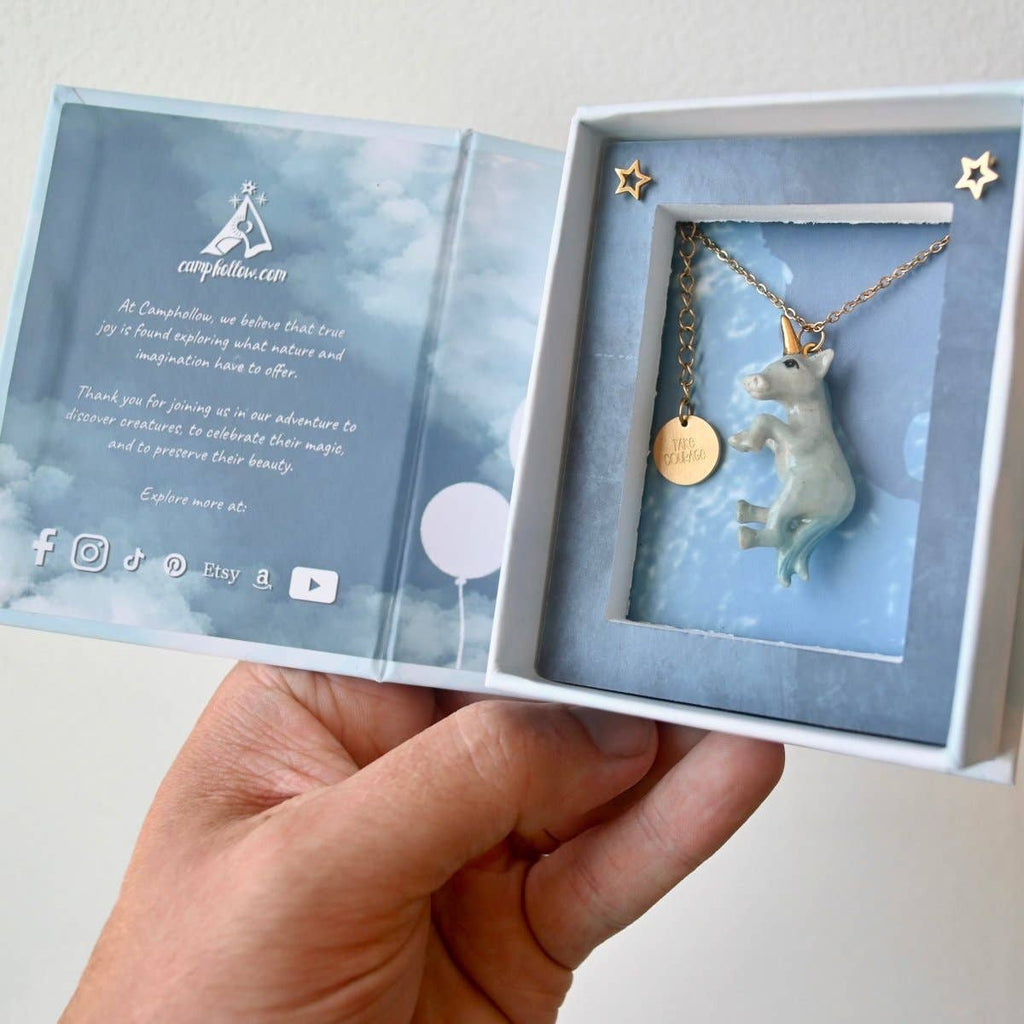 A hand holds an open jewelry box, unveiling the Galaxy Unicorn Necklace: a ceramic unicorn pendant on a gold chain. The box's interior is adorned with text and star decorations, and a round gold tag enhances the elegance of the porcelain piece. A cloudy sky design provides a charming backdrop.