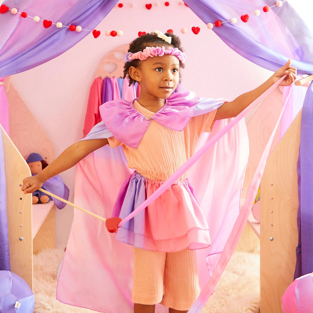 A young child stands in a playroom decorated with garlands of purple and pink fabric. They are dressed in a pink and purple fairy costume with a matching Sarah's Silk Blossom Garland Headband, perfect for dress-up play. The child holds a toy wand with a heart at the tip and looks to the side with a slight smile.