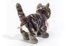 The Teddy Hermann Standing Tabby Cat Stuffed Animal is a plush toy that resembles a gray tabby cat with black stripes, standing on all fours and facing away. This cuddly cat features a short tail and appears soft and fluffy due to its high-quality plush material. Tags attached to the side and tail likely contain care instructions and brand information.