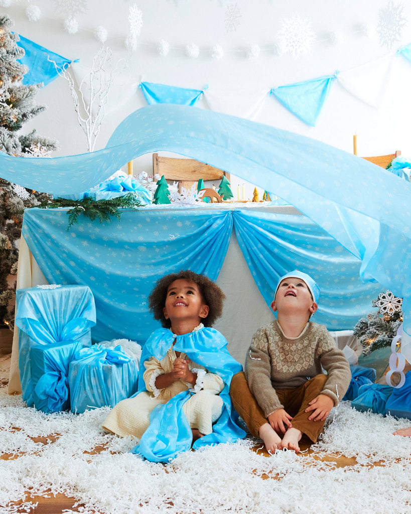 Two children sit on a snowy floor, captivated by the festive winter scene surrounding them. Surrounded by blue and white decorations, including snowflakes and Sarah's Silk Giant Snow Playsilk, one child dons a blue costume while the other wears a cozy sweater and pants, inviting open-ended play.