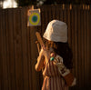 A young child with long hair, wearing a light-colored sun hat and a striped dress, is standing near a wooden fence. The child is holding a Wooden Bow & Arrows Set with Goal and has a quiver of arrows on their back, aiming at the target pinned to the fence. The scene, perfect for family fun, is bathed in warm sunlight.
