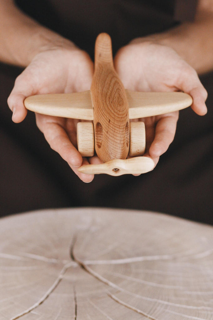 Hands holding a small wooden airplane above a wooden surface with a circular pattern. The airplane, crafted from light-colored wood and treated with natural linseed oil, boasts visible grain that enhances its rustic and handmade charm.