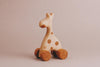 The Handmade Wooden Mini Giraffe Push Toy features a charming design with four small wheels and is made from light-colored wood. It showcases darker circular spots, adding elegance as it stands against a plain beige background, exuding timeless appeal.