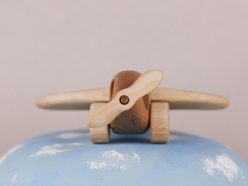 A Handmade Wooden Airplane, crafted with a propeller and enriched by natural linseed oil, is perched on a painted blue globe adorned with white clouds, set against a plain background.