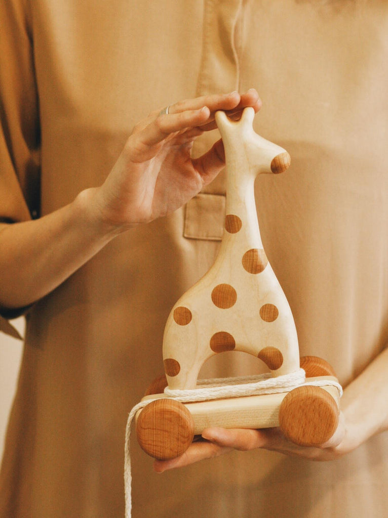 An individual wearing a beige shirt holds a Handmade Wooden Giraffe Pull Toy, adorned with delightful brown spots and equipped with a playful pull string.