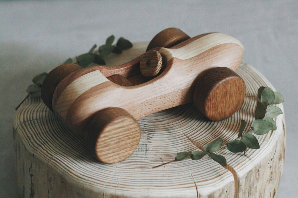 A Handmade Wooden Race Car sits atop a tree stump slice, surrounded by a few green leaves. The toy car has a simple, rustic design with ultra smooth curves and light and dark wood tones, finished with natural linseed oil.