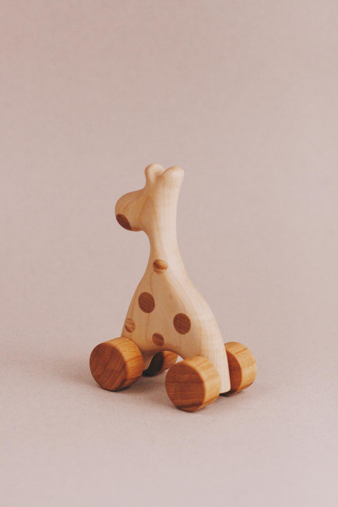 The Handmade Wooden Mini Giraffe Push Toy features a smooth finish and charming spotted details, mounted on four round wheels. It stands gracefully against a plain, neutral background.