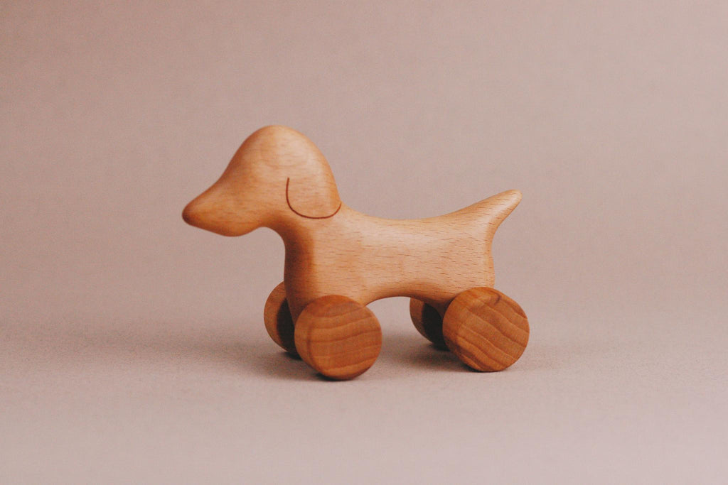 The Handmade Wooden Mini Dog Push Toy, crafted with smooth edges and a natural finish from maple and beech wood, stands against a plain beige background. Simple in design, this wooden toy dog is mounted on four wheels.