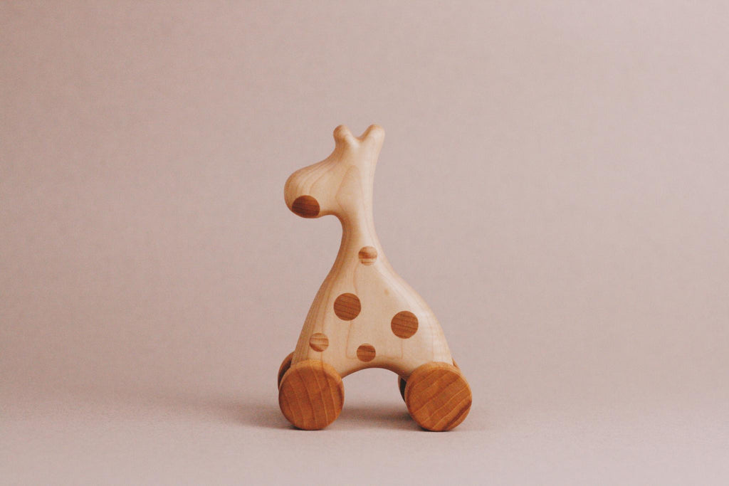 The Handmade Wooden Mini Giraffe Push Toy showcases wheels and painted spots on a minimalist beige background, crafted from maple and beech wood.